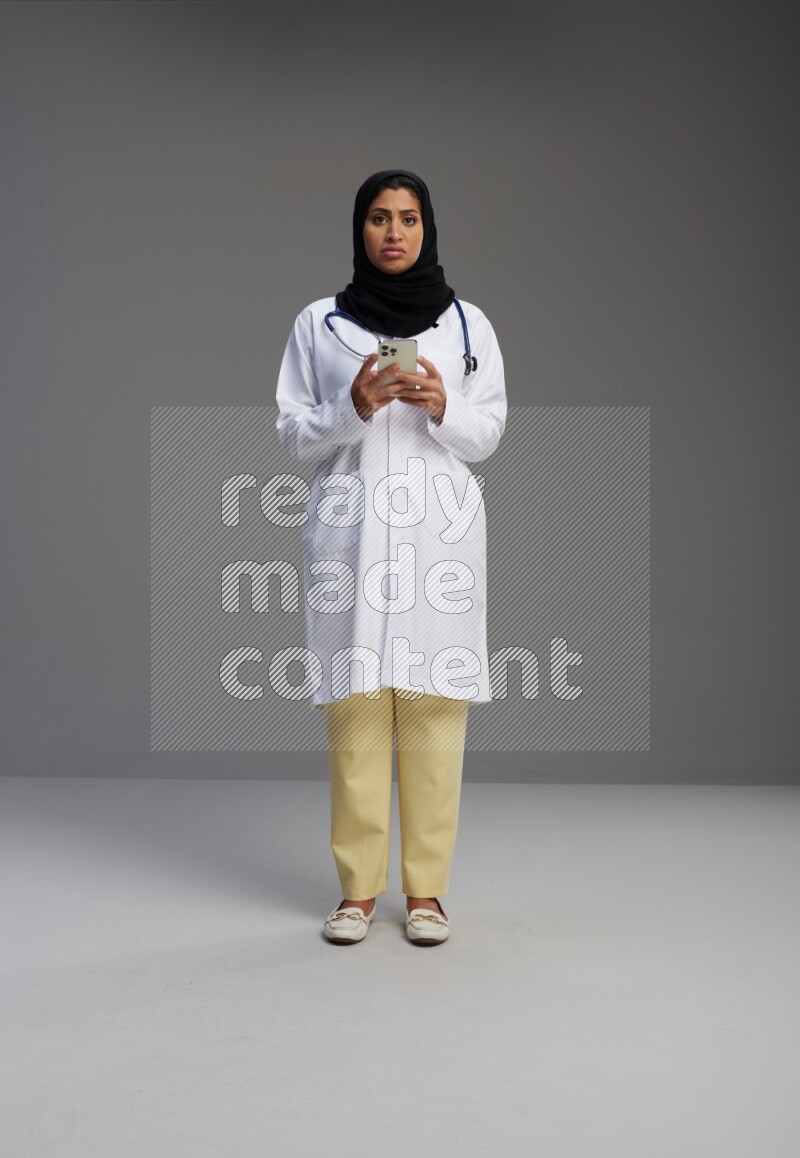 Saudi woman wearing lab coat with stethoscope standing texting on phone on Gray background