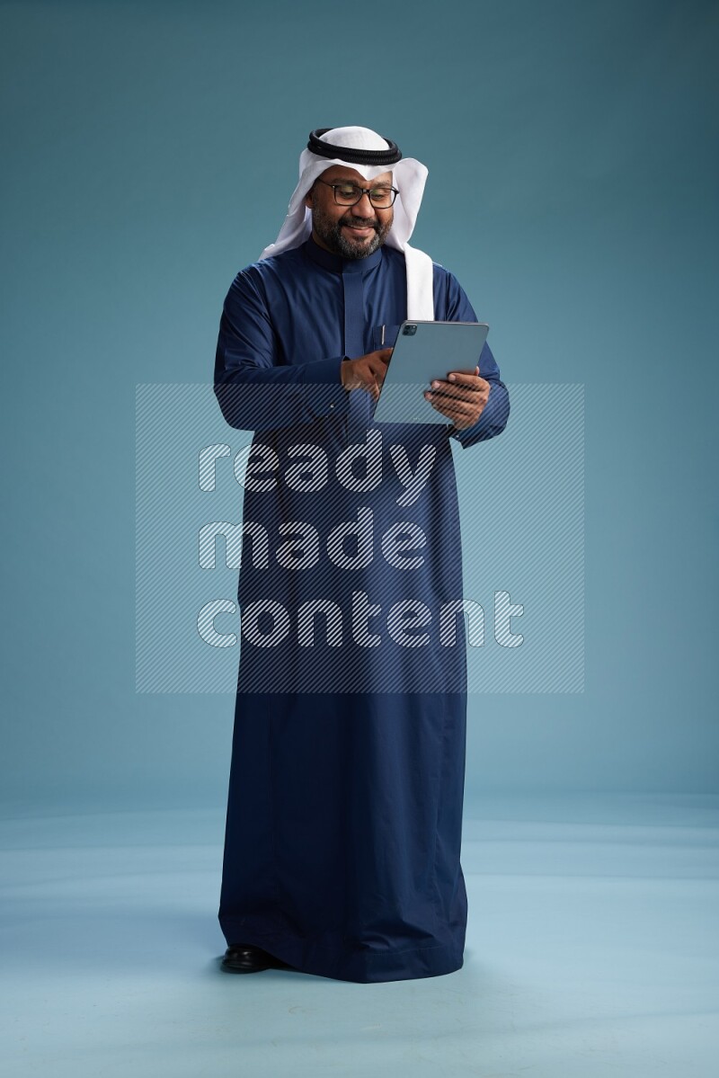 Saudi Man with shimag Standing holding ATM card while working on tablet on blue background