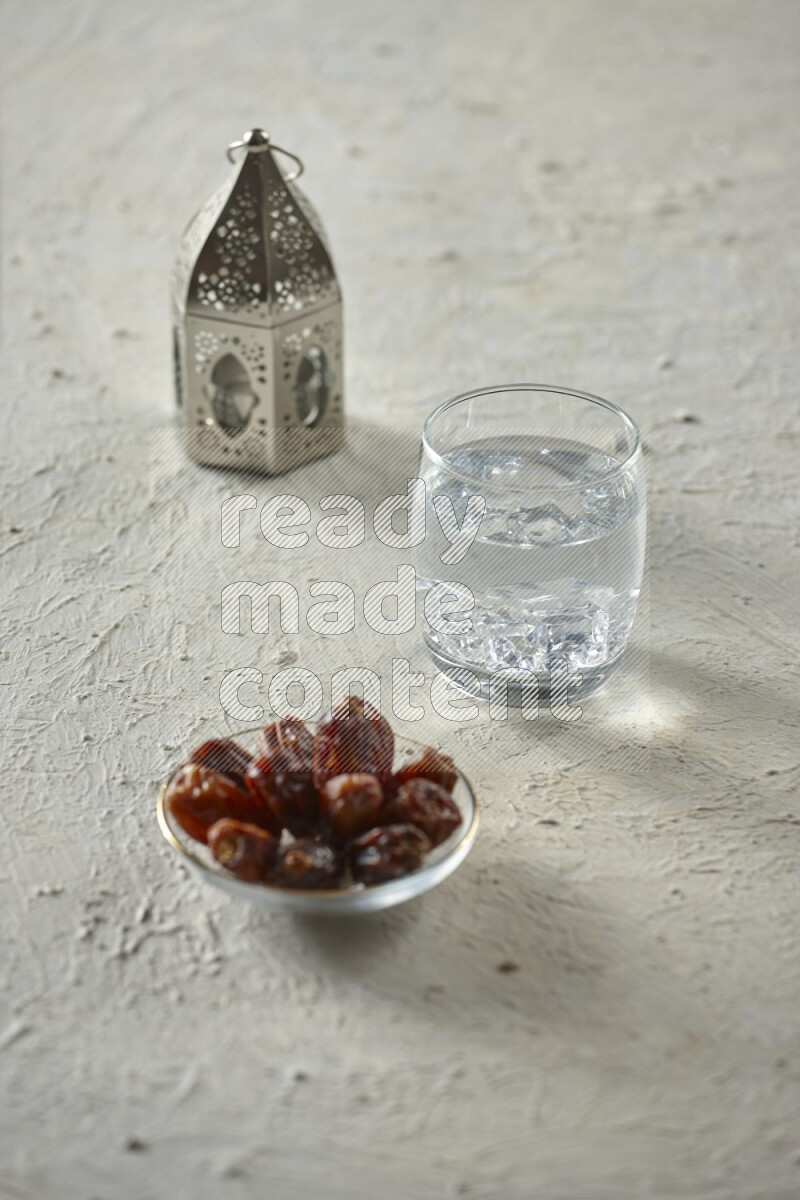 A silver lantern with different drinks, dates, nuts, prayer beads and quran on textured white background