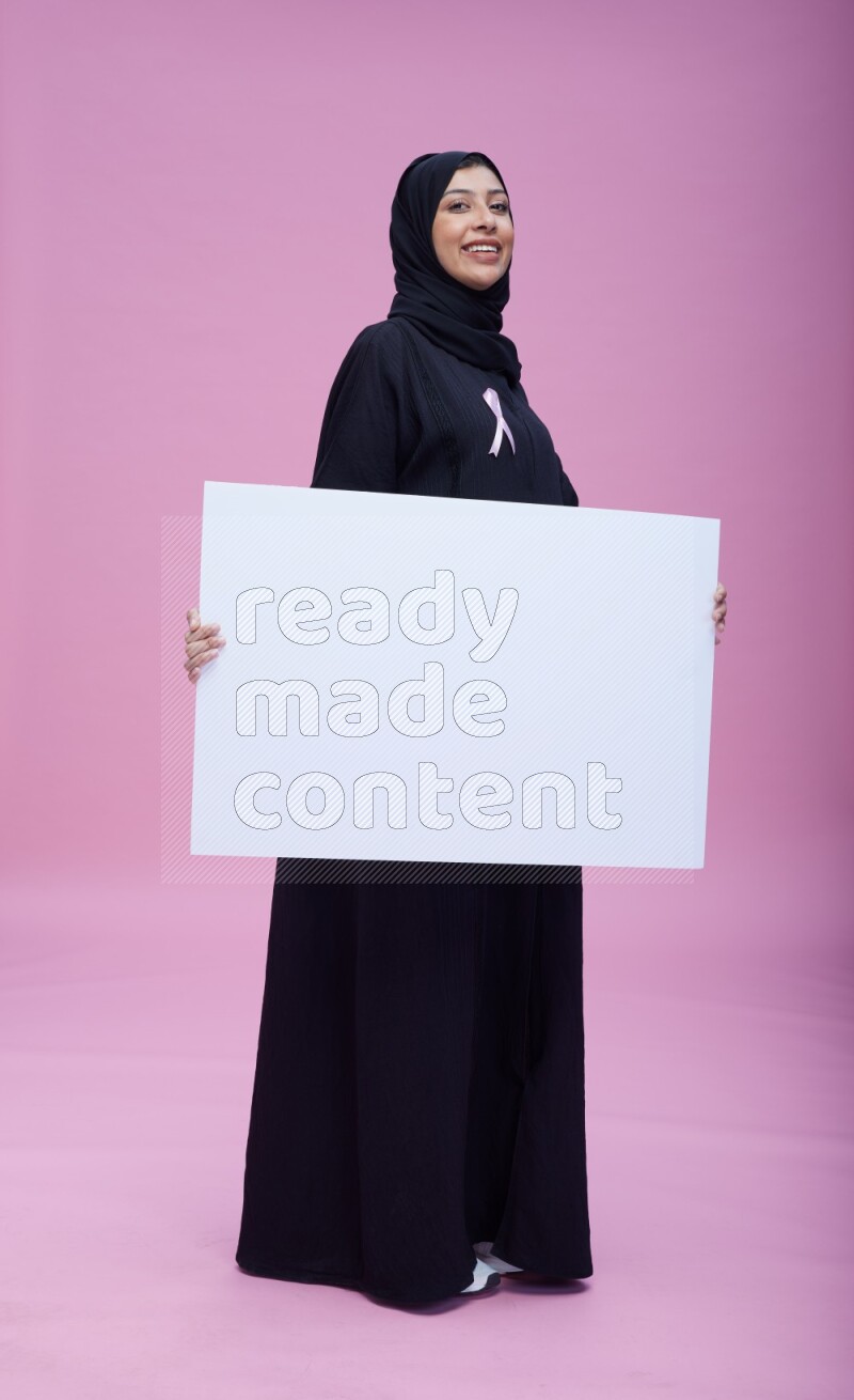 Saudi woman wearing pink ribbon on Abaya standing holding board on pink background