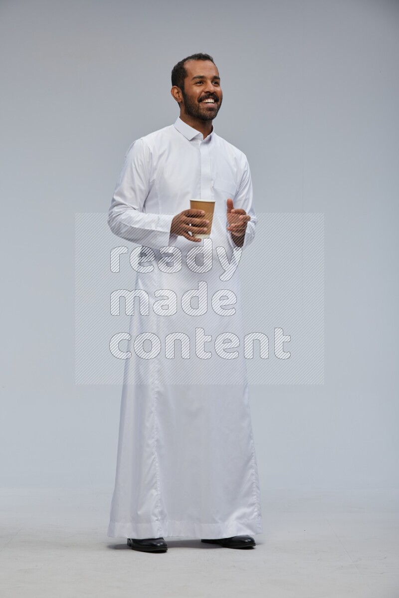 Saudi man wearing thob standing holding paper cup on gray background