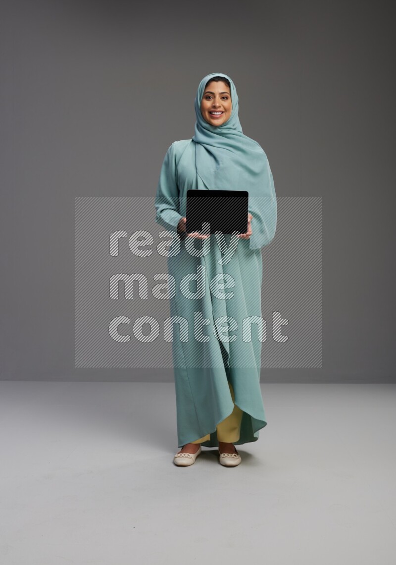 Saudi Woman wearing Abaya standing showing tablet to camera on Gray background