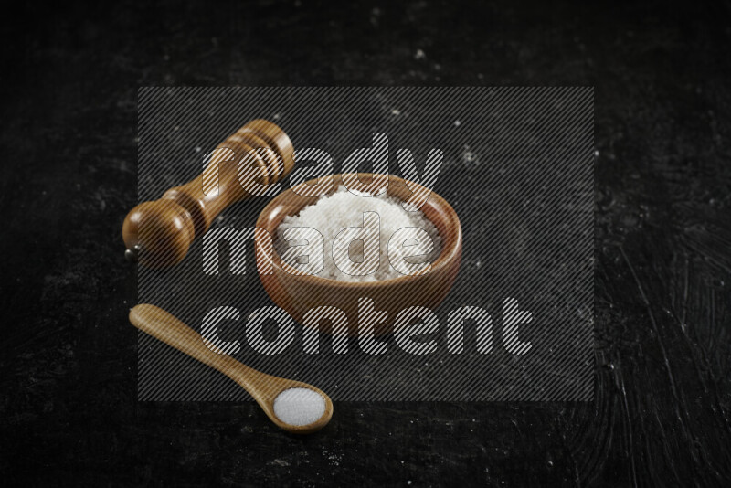 A wooden bowl and spoon filled with white sea salt and wooden grinder beside them on black background