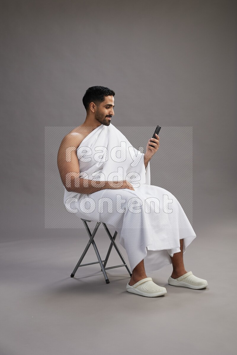 A man wearing Ehram sitting on chair texting on phone on gray background