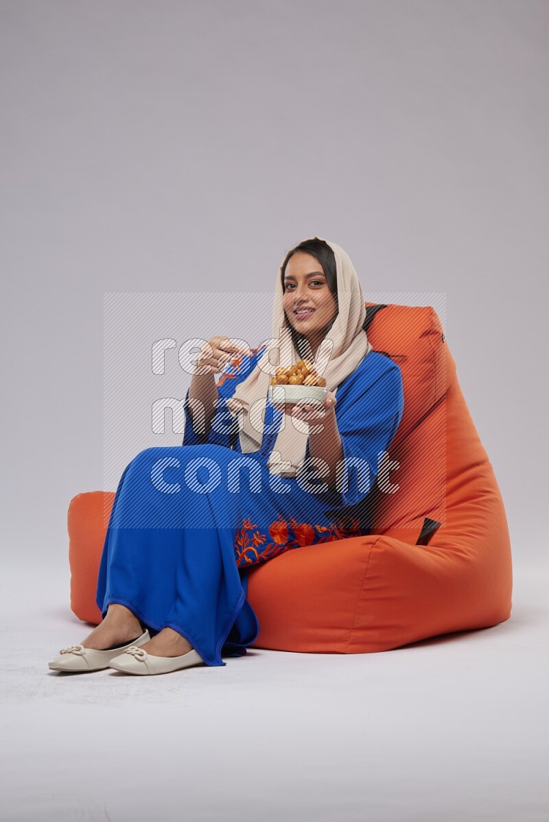 A Woman sitting on an orange beanbag wearing Jalabeya holding a plate of luqaimat