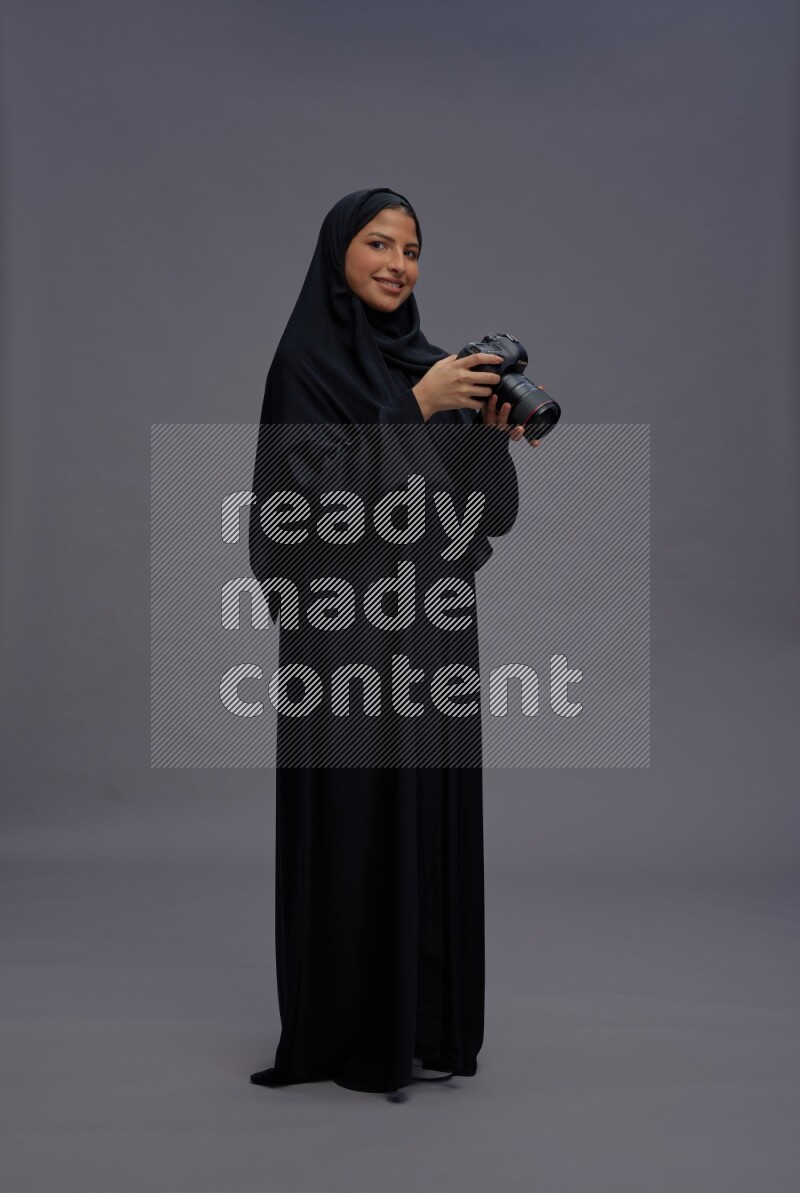 Saudi woman wearing Abaya standing holding Camera on gray background