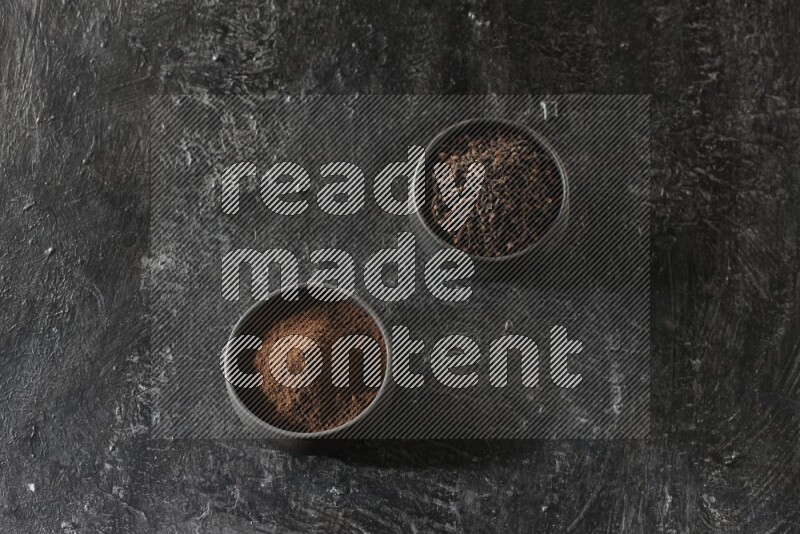 2 Black pottery bowls full of cloves and the other full of cloves powder on textured black flooring