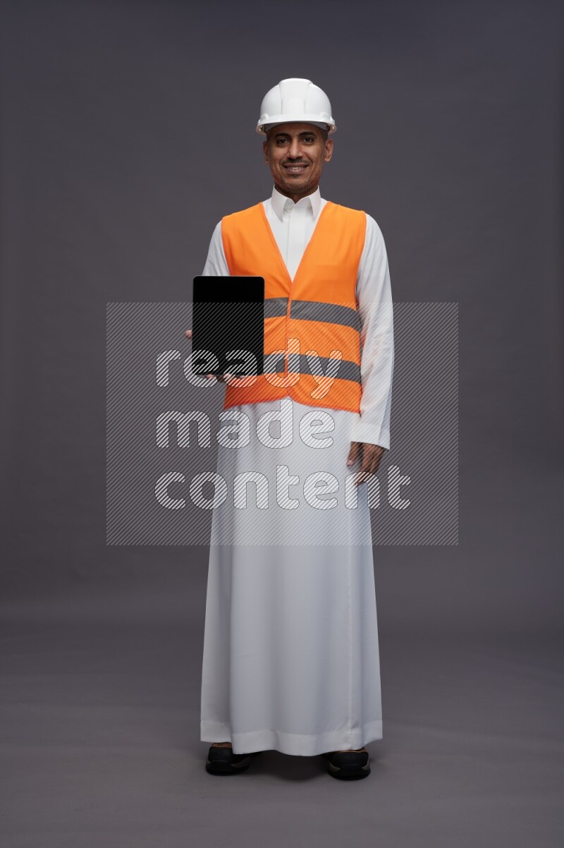 Saudi man wearing thob with engineer vest standing showing tablet to camera on gray background