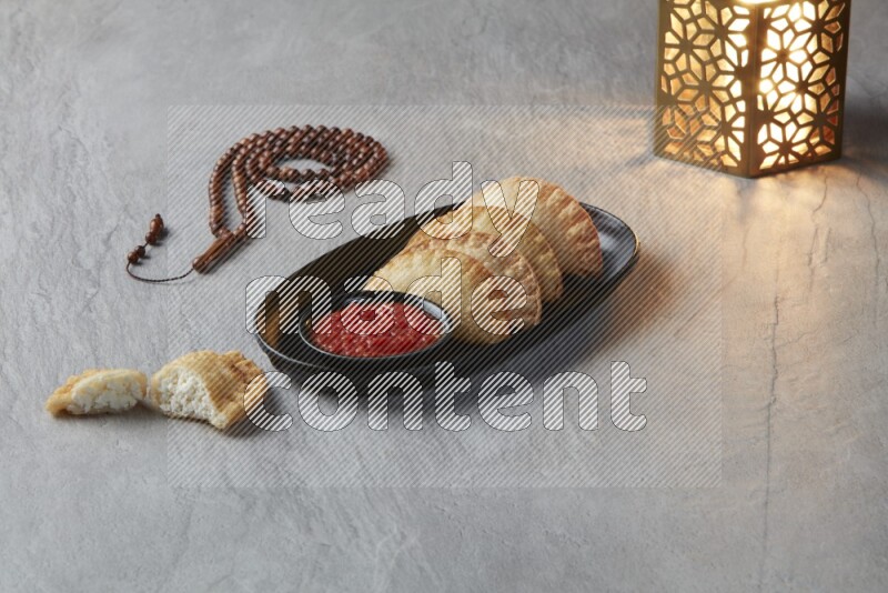Four fried sambosas in an oval shaped black plate, beside a cut cheese sambosa, a brown misbaha and a golden lantern on a gray background