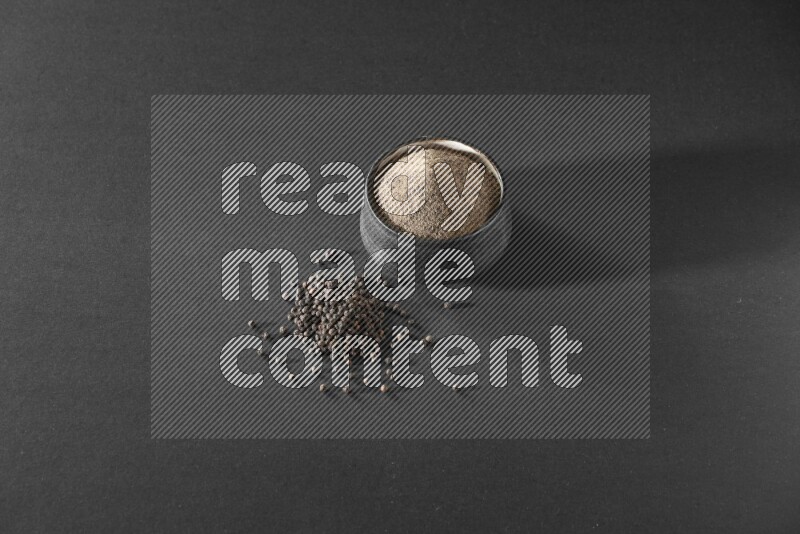 A black pottery bowl full of black pepper powder and black pepper beads on the floor on black flooring