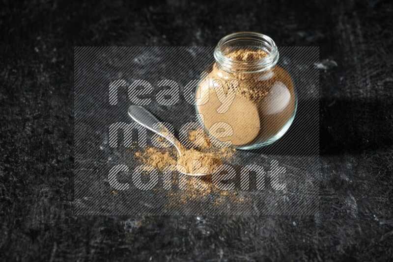 A glass spice jar and metal spoon full of allspice powder on a textured black flooring