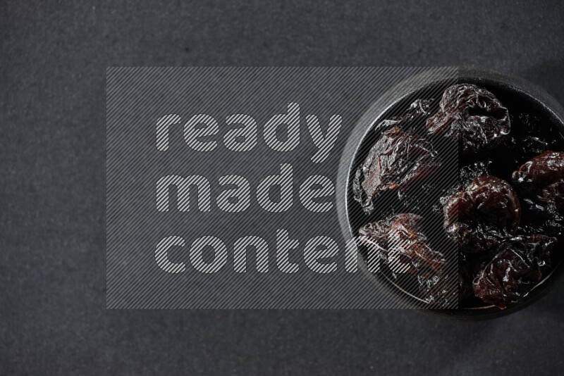 A black pottery bowl full of dried plums on a black background in different angles