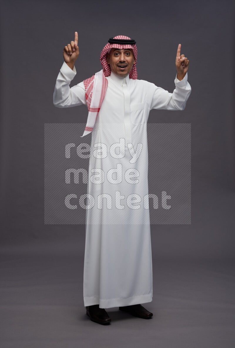 Saudi man wearing thob and shomag standing interacting with the camera on gray background