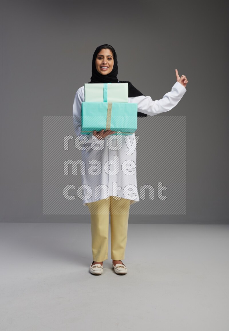 Saudi woman wearing lab coat with stethoscope standing holding gift box on Gray background