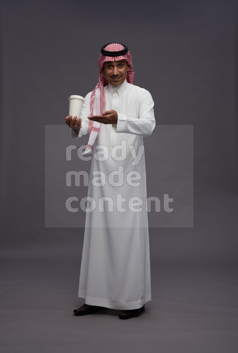 Saudi man wearing thob and shomag standing holding paper cup on gray background