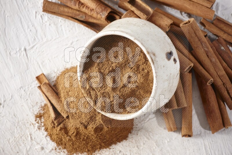 Ceramic beige bowl over filled with cinnamon powder and cinnamon sticks around the bowl on a textured white background in different angles