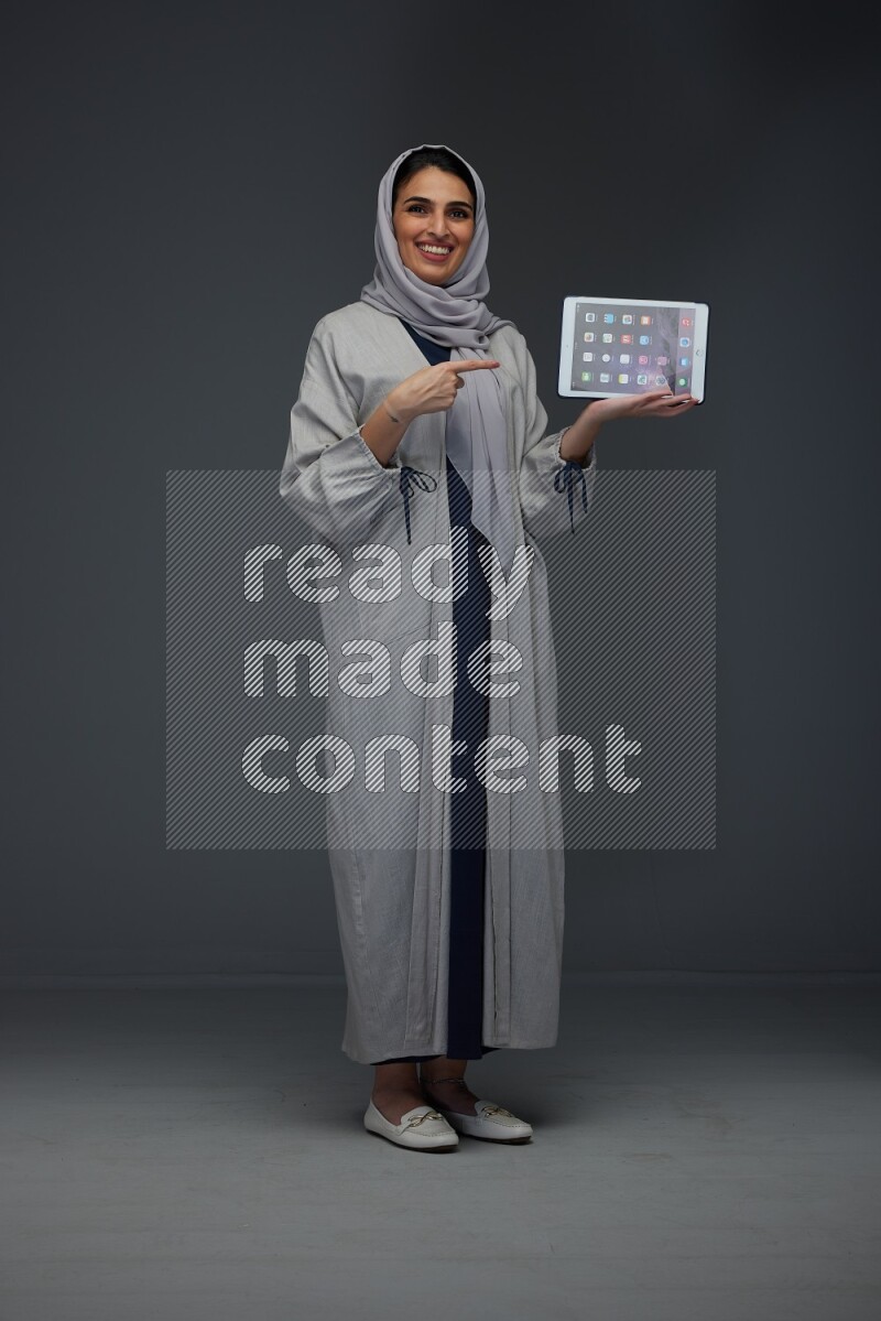 A Saudi woman wearing a light gray Abaya and head scarf standing and showing the phone's screen eye level on a grey background