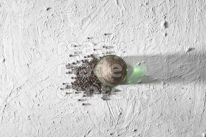 A glass cup full of black pepper powder and black pepper spread on a textured white flooring