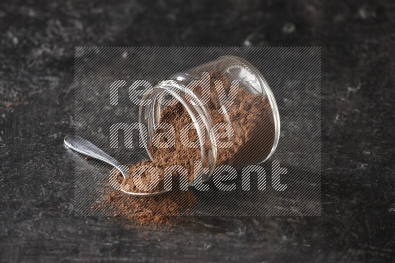 A flipped glass jar and metal spoon full of cloves powder with cloves spread on a textured black flooring