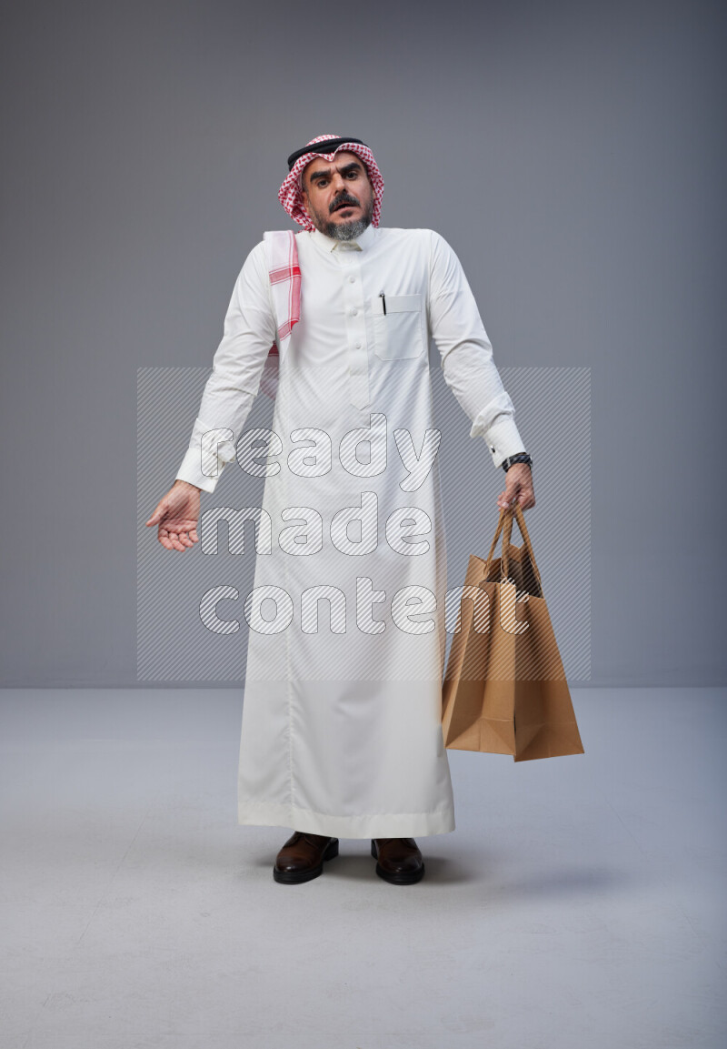 Saudi man Wearing Thob and red Shomag standing holding shopping bag on Gray background