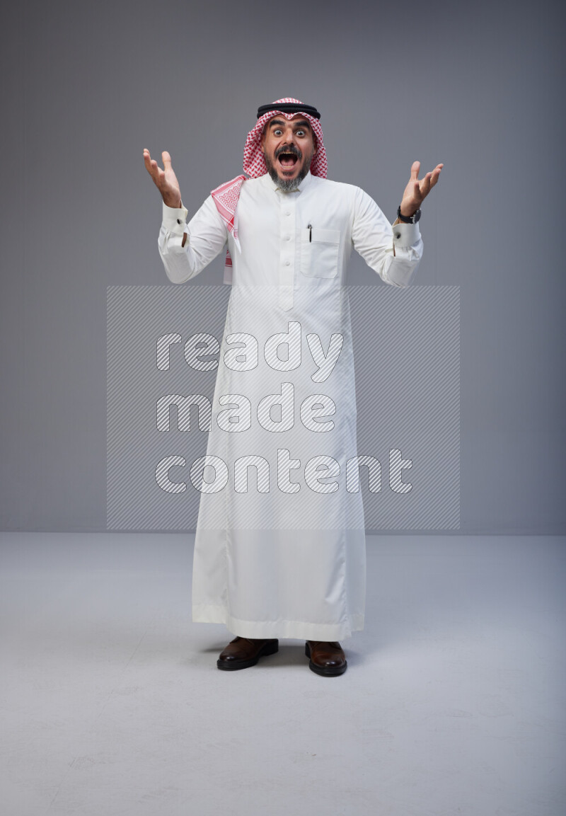 Saudi man Wearing Thob and red white Shomag standing interacting with the camera on Gray background