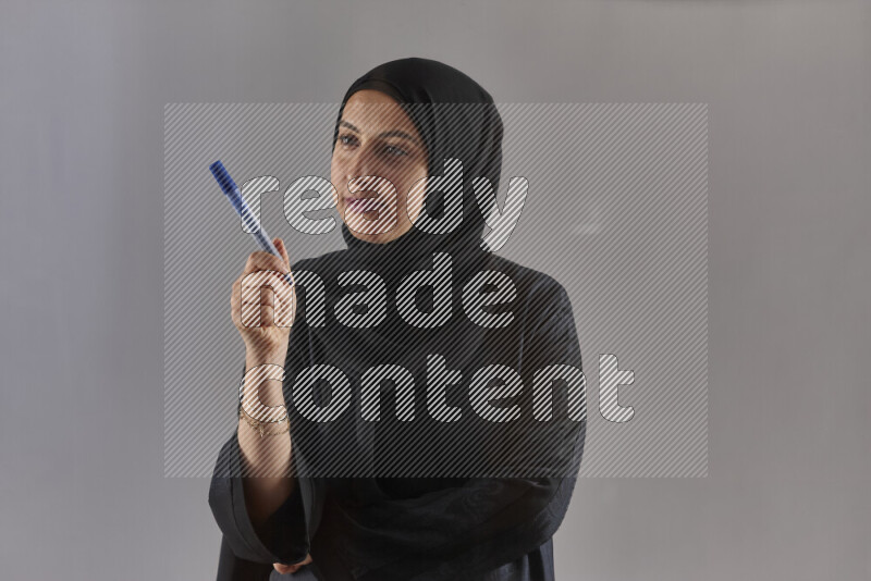 Woman in black abaya and hijab holding a marker pen to write on a transparent board with different reactions and expressions on grey background