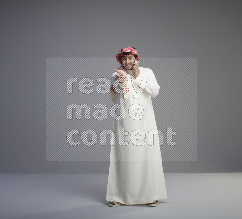 A Saudi man standing wearing thob and red shomag talking on phone on gray background