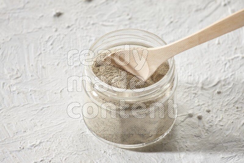 A glass jar and wooden spoon full of white pepper powder on textured white flooring
