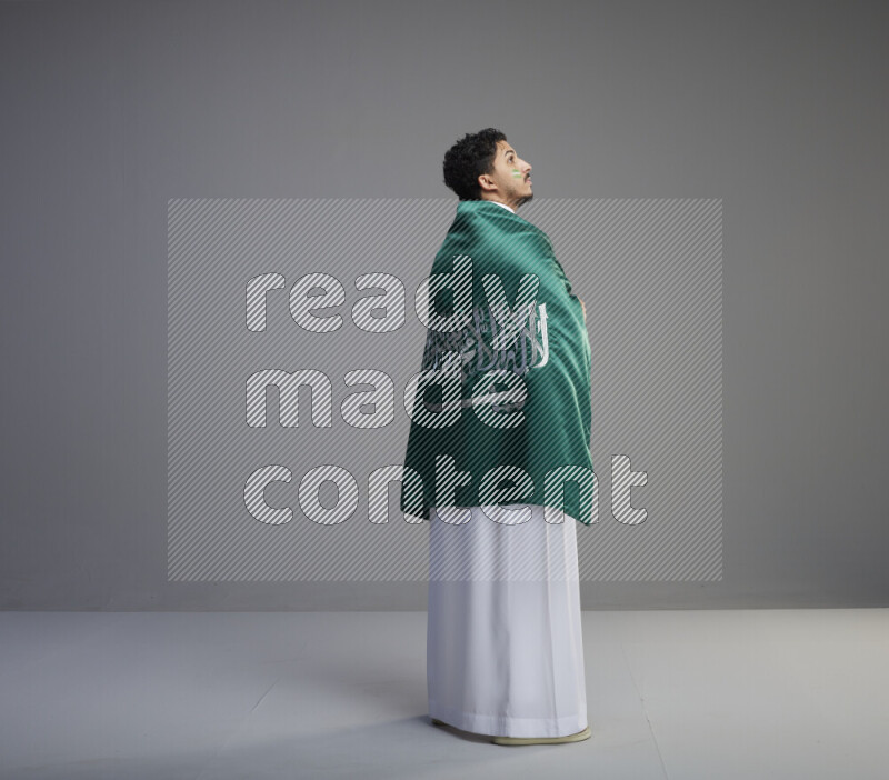A Saudi man standing wearing thob with face painting wrapping big Saudi flag on gray background