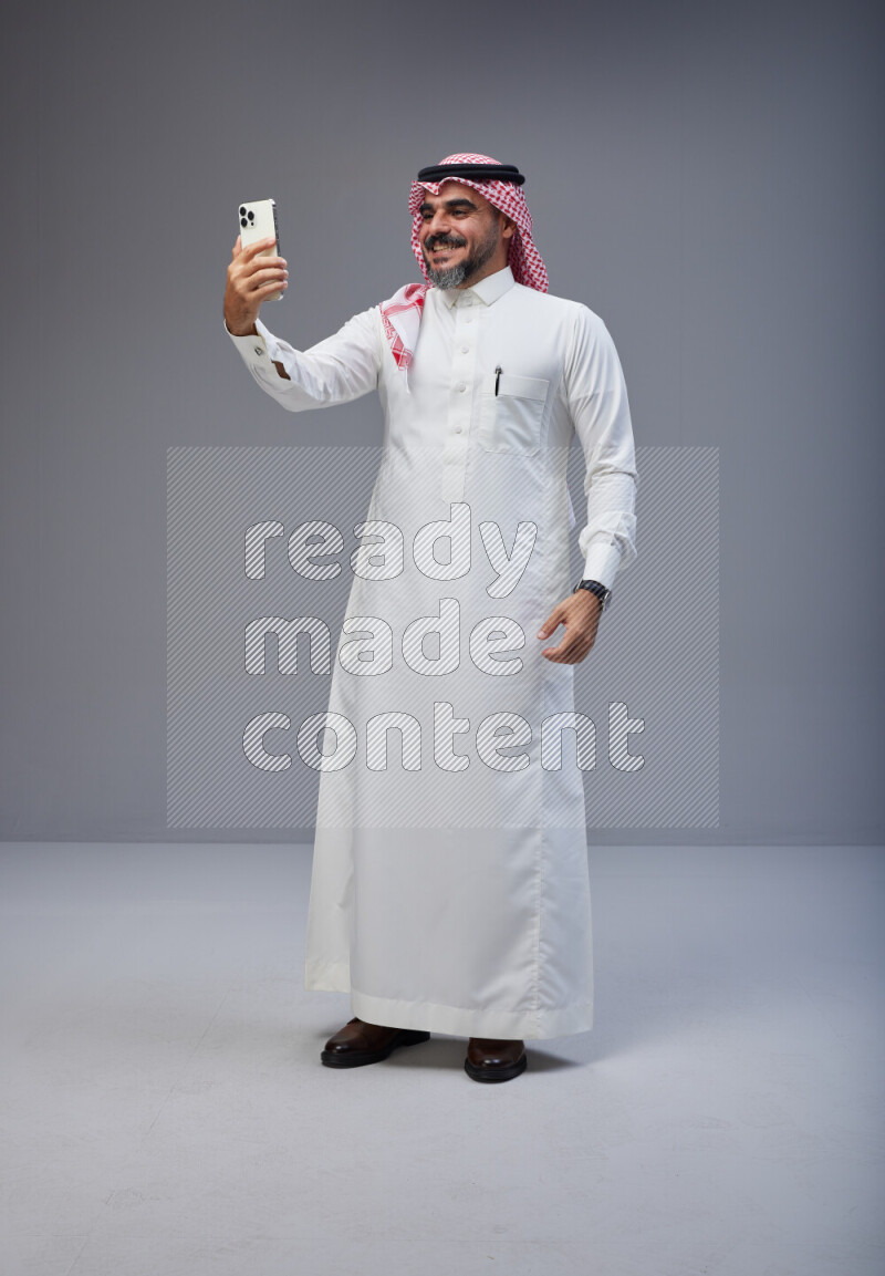 Saudi man Wearing Thob and red Shomag standing taking selfie on Gray background