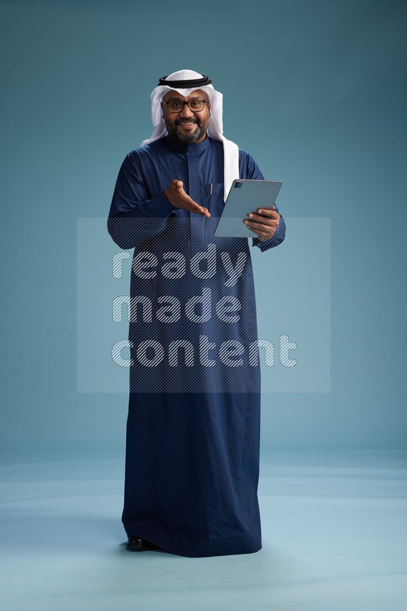 Saudi Man with shimag Standing holding ATM card while working on tablet on blue background