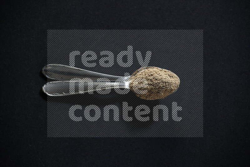 2 Metal spoons full of cardamom powder on black flooring