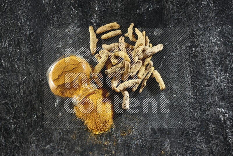 وعاء زجاجي للتوابل ممتلئ ببودرة الكركم مقلوب والبودرة تناثرت منه مع أصابع الكركم المجففة على خلفية سوداء