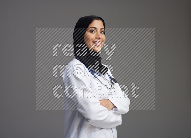 Saudi woman wearing lab coat with stethoscope standing with crossed arms on Gray background