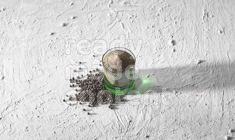 A glass cup full of black pepper powder and black pepper spread on a textured white flooring