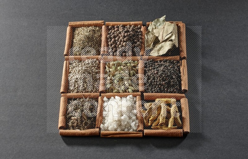 9 squares of cinnamon sticks full of cardamom in the middle surrounded by turmeric fingers, black pepper, bay laurel leaves, allspice, cumin, white pepper, dried basil and salt on black flooring