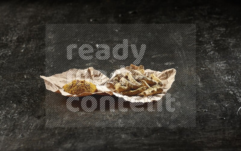 Turmeric powder and dried turmeric whole fingers in 2 crumpled pieces of paper on textured black flooring