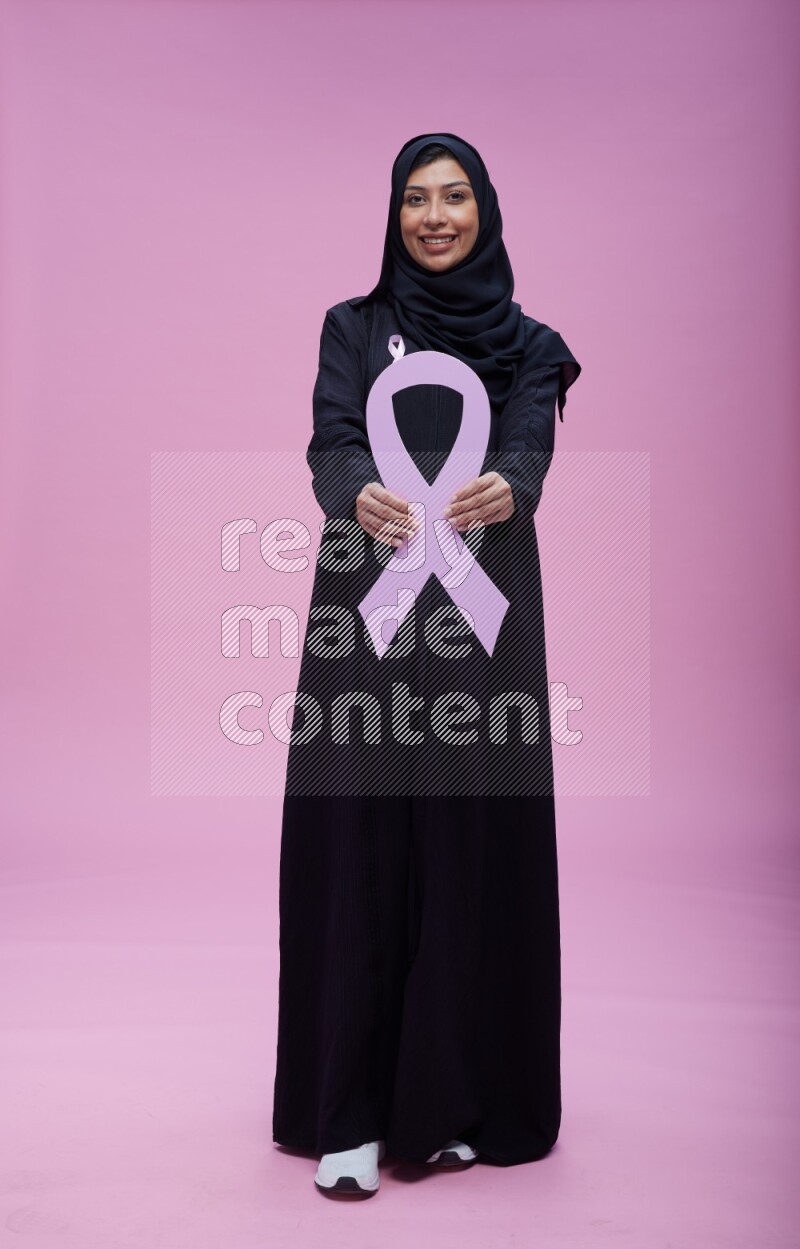 Saudi woman wearing pink ribbon on abaya standing holding awareness ribbon on plain pink background