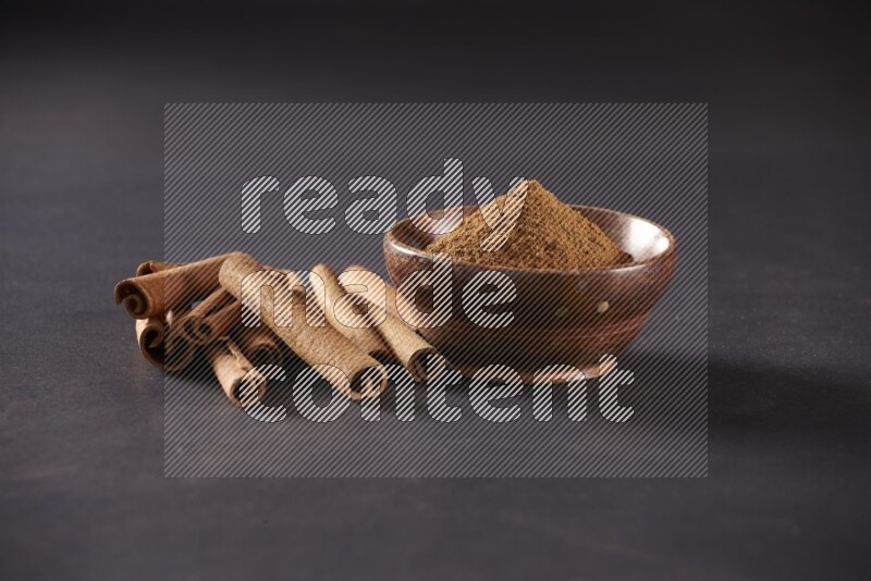 Cinnamon sticks stacked beside a wooden bowl full of cinnamon powder on black background