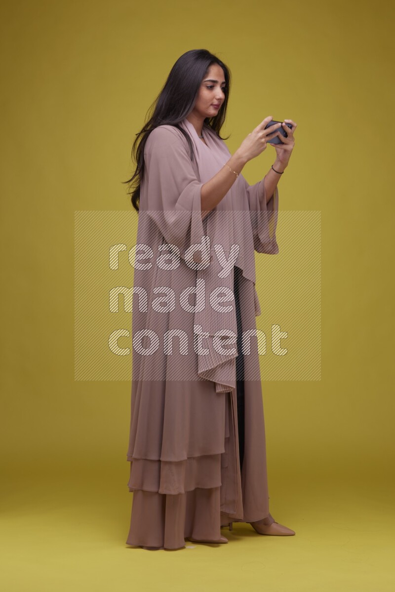 A woman Playing a Game on her smartphone on a Yellow Background wearing Brown Abaya