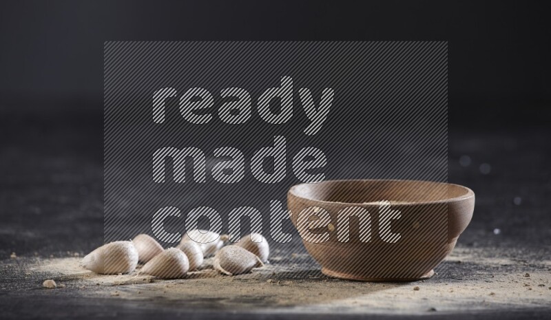 وعاء خشبي ممتلئ ببودرة الثوم مع بعض فصوص الثوم بجانبه علي خلفية سوداء