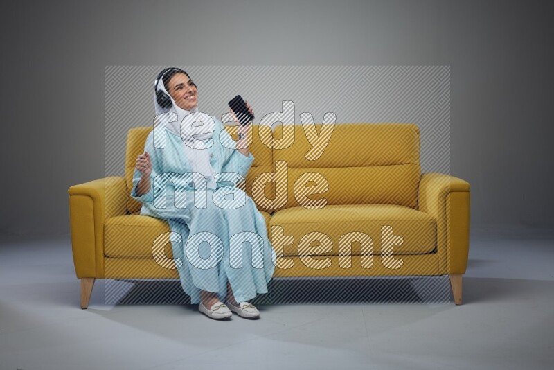 A Saudi woman wearing a light blue Abaya and a white head scarf sitting on a yellow sofa and holding an electronic while wearing headphone eye level on a grey background