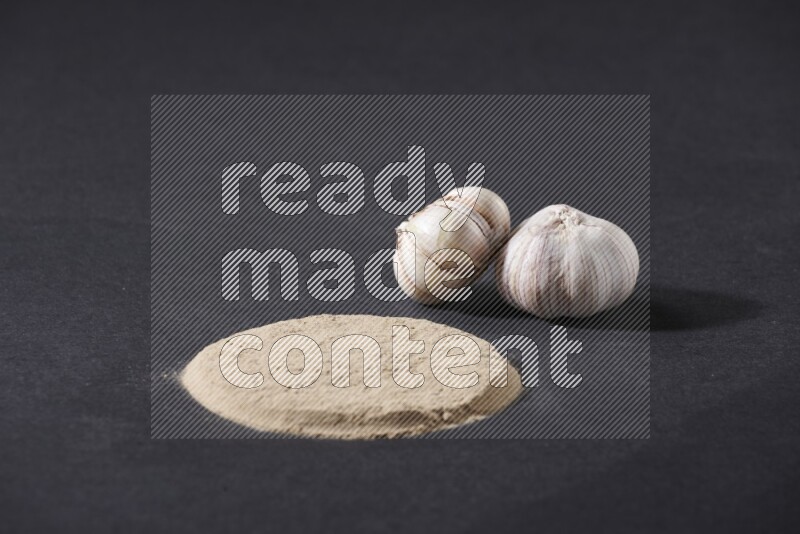 Garlic powder in a circle shape and beside it 2 garlic bulbs on a black flooring