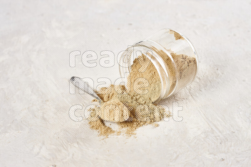 A glass jar full of ground ginger powder flipped with some spilling powder on white background