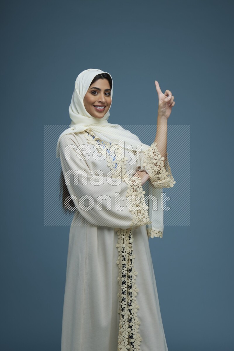 A Saudi woman pointing in a blue background wearing an off-white Abaya Hijab
