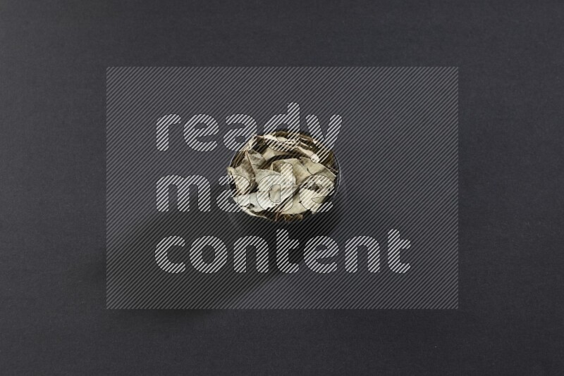A black pottery bowl full of dried bay leaves on black flooring