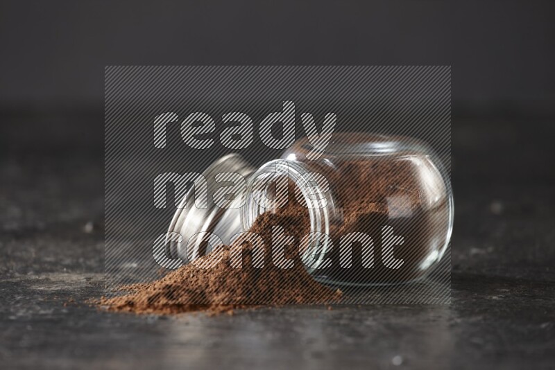 A flipped glass spice jar full of cloves powder and powder came out of it on textured black flooring