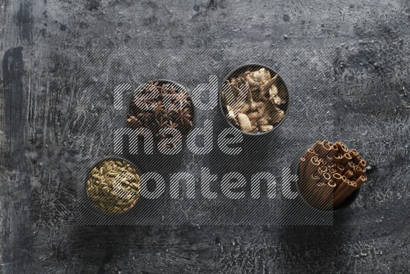 Ginger, Cardamom, Star anise and cinnamon sticks in 4 bowls on a textured black background