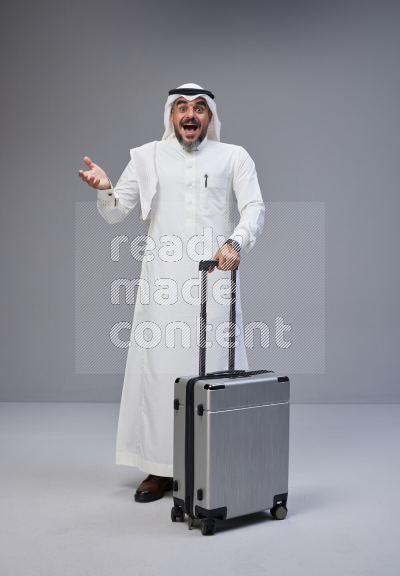 Saudi man wearing Thob and white Shomag standing holding Travel bag on Gray background