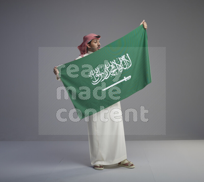 A saudi man standing wearing thob and red shomag holding big saudi flag on gray background